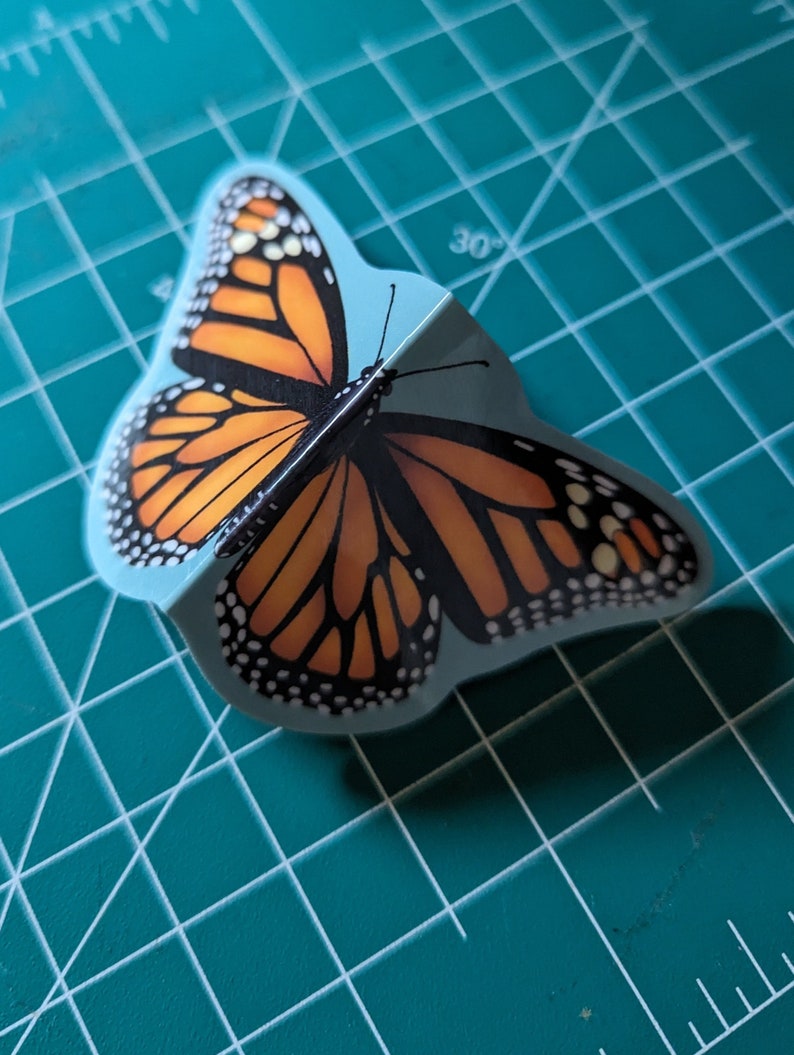 May include: A three-dimensional monarch butterfly sticker featuring vibrant orange and black wings with white spots along the edges. The sticker is folded along the center body, creating a raised effect that mimics the appearance of a butterfly in flight.