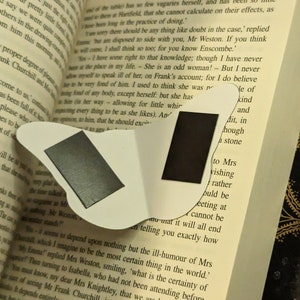 May include: A white butterfly-shaped magnetic bookmark featuring two rectangular black magnetic strips on its surface, positioned over an open book with printed text visible on the pages, including the word Emma at the top of the page.