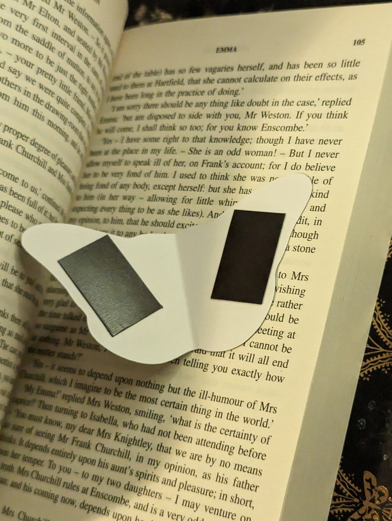 May include: A white butterfly-shaped magnetic bookmark featuring two rectangular black magnetic strips on its surface, positioned over an open book with printed text visible on the pages, including the word Emma at the top of the page.