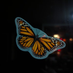 May include: A monarch butterfly sticker featuring vibrant orange and black wings with white spots along the edges. The butterfly is depicted with a light blue border against a dark background, showcasing detailed wing patterns and thin antennae.