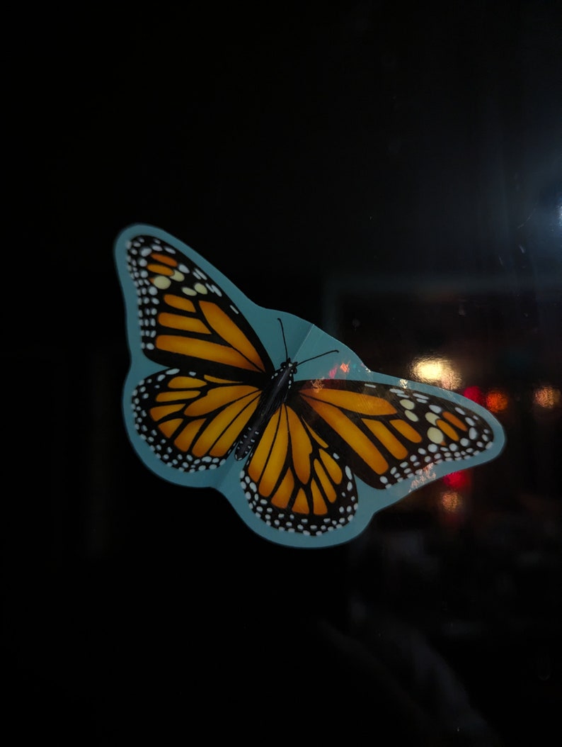 May include: A monarch butterfly sticker featuring vibrant orange and black wings with white spots along the edges. The butterfly is depicted with a light blue border against a dark background, showcasing detailed wing patterns and thin antennae.