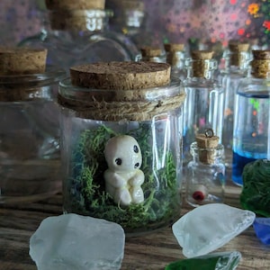 May include: A selection of glass jars with cork stoppers. One jar contains a small, white figurine in green moss. Other jars hold coloured liquids and decorative elements, including sea glass.