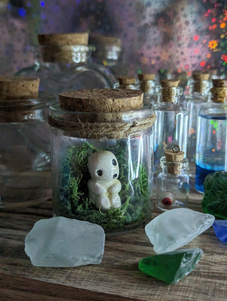 May include: A selection of glass jars with cork stoppers. One jar contains a small, white figurine in green moss. Other jars hold coloured liquids and decorative elements, including sea glass.