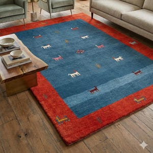 Puede incluir: Una alfombra rectangular con un centro azul y un borde rojo. El campo azul presenta pequeñas figuras de animales estilizadas en blanco, rojo y marrón. La alfombra está colocada sobre un suelo de madera, con muebles al fondo.