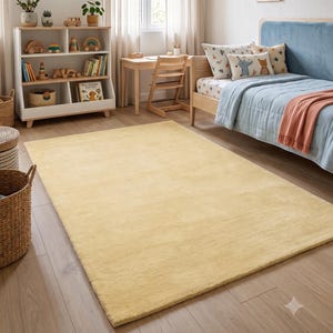 May include: A rectangular, solid light yellow area rug with a soft, uniform texture. The rug is positioned on a light-coloured wooden floor in a room featuring a bed with blue bedding, a wooden bookshelf, a small desk, and wall art.