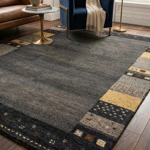 Puede incluir: Una alfombra rectangular grande con un campo central gris oscuro y un borde estampado. El borde presenta secciones negras, amarillas y grises con diseños geométricos y abstractos. La alfombra está sobre un suelo de madera.
