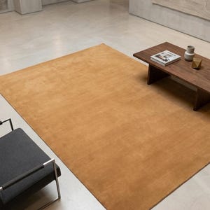 May include: A large, rectangular, solid mustard-yellow area rug with a smooth, uniform texture. The rug is positioned on a light-coloured floor, partially extending under a dark wood coffee table and near a dark grey chair with a metal frame.