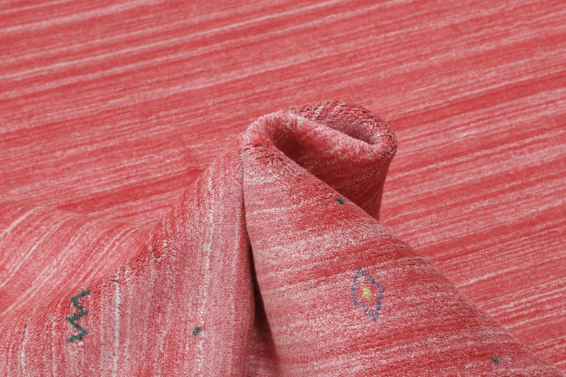 Puede incluir: Una alfombra roja con un patr&oacute;n de rayas horizontales texturizadas en varios tonos de rojo y blanco. La superficie presenta peque&ntilde;os motivos geom&eacute;tricos bordados dispersos en verde, azul y amarillo, incluyendo formas de zigzag y diamantes.