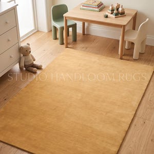 Peut inclure: Un tapis rectangulaire de couleur jaune moutarde uni avec une texture lisse à poils ras. Le tapis est posé sur un parquet en bois clair dans une pièce avec une table en bois, deux petites chaises, un ours en peluche et une commode. Texte STUDIO HANDLOOM RUGS.