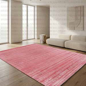 Puede incluir: Una alfombra rectangular que presenta una paleta de colores rosa vibrante con sutiles estr&iacute;as horizontales blancas y una textura desgastada en toda la superficie. La alfombra est&aacute; colocada sobre un suelo de madera en una habitaci&oacute;n clara.