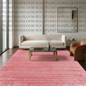 Puede incluir: Una alfombra rectangular de un tono rosa vibrante que presenta una textura sutil de rayas horizontales con tenues acentos oscuros dispersos por todo el tejido. La alfombra cubre el espacio del suelo frente a un sof&aacute; color crema y una mesa de centro verde.