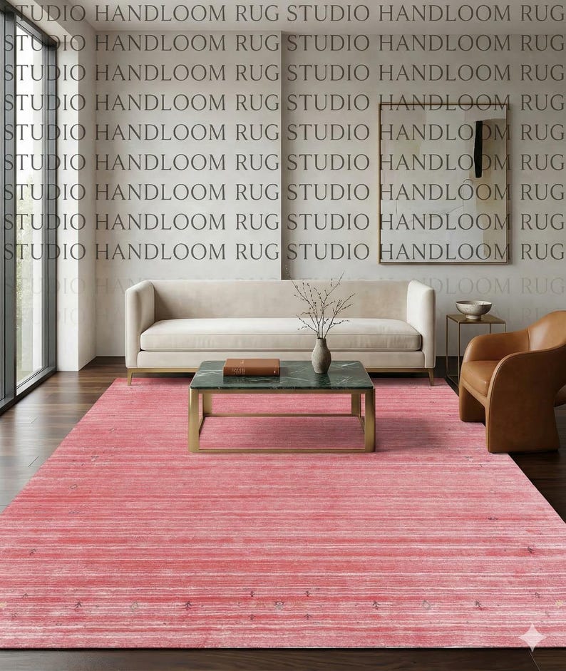 Puede incluir: Una alfombra rectangular de un tono rosa vibrante que presenta una textura sutil de rayas horizontales con tenues acentos oscuros dispersos por todo el tejido. La alfombra cubre el espacio del suelo frente a un sof&aacute; color crema y una mesa de centro verde.