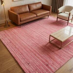 Puede incluir: Una alfombra rectangular grande en tonos rosa y blanco, con un patr&oacute;n lineal. La alfombra est&aacute; colocada en un sal&oacute;n, junto a un sof&aacute; de cuero marr&oacute;n, un sill&oacute;n blanco y una mesa de centro con tapa de m&aacute;rmol.