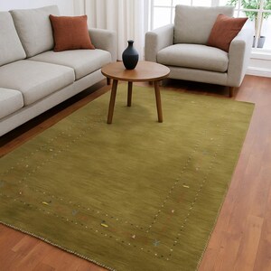 Pode incluir: Um tapete retangular verde-oliva com uma borda estampada, colocado sobre um piso de madeira. O tapete está em uma sala de estar com um sofá, uma poltrona e uma mesa de centro redonda de madeira. Duas almofadas cor de ferrugem estão no sofá e na poltrona.