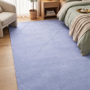 May include: A rectangular area rug in a solid light periwinkle blue color with a soft, textured pile surface. The rug features a repeating watermark text that reads STUDIO HANDLOOM RUGS across the surface of the fabric in a light grey tone.