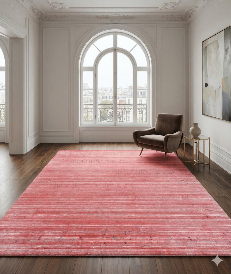Puede incluir: Una alfombra rectangular de un vibrante tono rojo coral que presenta un sutil patr&oacute;n de rayas horizontales con tenues motas oscuras dispersas por todo el tejido. La alfombra est&aacute; colocada sobre un suelo de madera oscura en una habitaci&oacute;n.