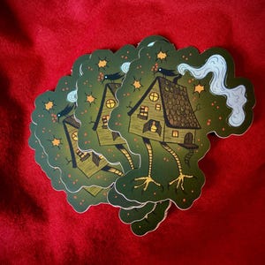 May include: A stack of green stickers featuring a cartoon illustration of a house with a chimney, a roof, and windows. The house is on long, spindly legs and has a bird perched on the roof. The background is a dark green with stars and red berries.