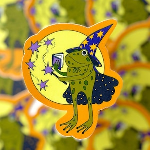 Op de afbeelding: Een grillige sticker met een groene kikker-tovenaar met een blauwe en gele hoed en cape. De kikker houdt een boek en een toverstaf vast, omringd door paarse sterren, tegen een gele cirkel en een oranje rand. De sticker heeft een cartoonstijl.