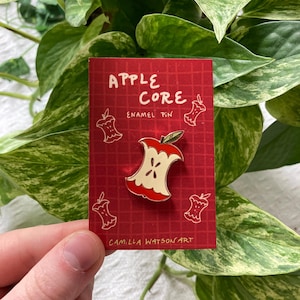 May include: A red enamel pin in the shape of an apple core, with a white interior and a green leaf. The pin is on a red card with the text "APPLE CORE ENAMEL PIN" and the artist's name. The background is a green leafy plant.