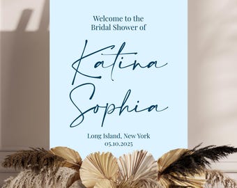 May include: A light blue welcome sign for a bridal shower, with the text "Welcome to the Bridal Shower of Katina Sophia" in elegant script. The sign also includes the location and date, and is decorated with dried flowers.