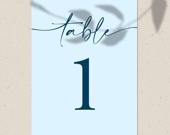 May include: A light blue table number card with the word "table" in a cursive font and the number "1" in a bold font. The card is rectangular and has a shadow of leaves in the background.