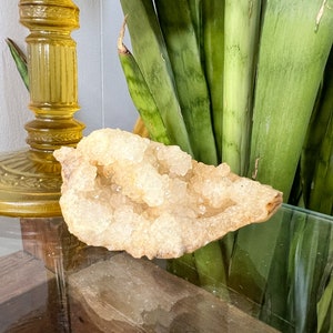 May include: A large, light brown, crystal cluster with a rough, natural texture. The cluster is made up of many small, clear crystals that are clustered together. The crystal cluster is sitting on a glass surface.