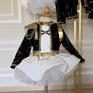 May include: A child's costume with a black sequined jacket, gold trim, and a white tutu skirt. The jacket features gold epaulets, chains, and a tassel. The outfit is displayed on a mannequin.