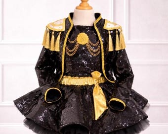 May include: A black sequined dress with a gold-trimmed jacket and matching gold accents. The jacket features gold tassels and shoulder details. The dress has a flared skirt with a black tulle hem and a gold bow at the waist. The outfit is displayed on a mannequin.
