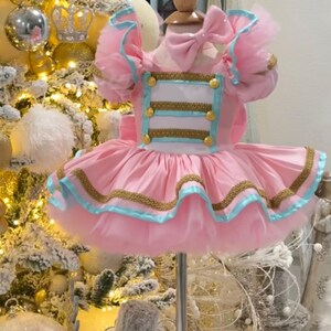 May include: A pink tutu dress with gold trim and light blue accents. The bodice features gold buttons and trim, with a matching bow at the neckline. The dress is displayed on a mannequin, with a Christmas tree in the background.