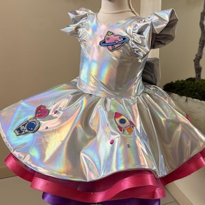 May include: A silver, iridescent dress with a space theme. The dress features a round neckline, short ruffled sleeves, and a full skirt. Appliqués include a heart-shaped planet, a rocket, and a telescope. The skirt has layers of pink and purple trim.