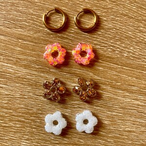Könnte beinhalten: Eine Auswahl an Ohrringen in verschiedenen Stilen. Zwei Paar goldene Creolen, zwei Paar blütenförmige Ohrringe mit rosa und orangefarbenem Glitzer, zwei Paar blütenförmige Ohrringe mit braunen und goldenen Strasssteinen und zwei Paar weiße Blütenohrringe.