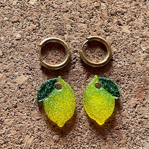 Könnte beinhalten: Ein Paar goldfarbene Creolen und zwei zitronenförmige Ohrringe. Die Zitronenohrringe sind gelb mit grünem Glitzer und einem grünen Blatt. Auf einer braunen, strukturierten Oberfläche.
