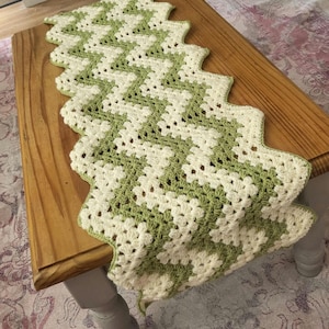 May include: A handmade crochet table runner with a zig-zag design. The runner has alternating rows of cream and light green. The edges are scalloped. The runner is displayed on a wooden table.