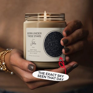 May include: A beige candle in a glass jar with a lit wick. The label reads "BORN UNDER THESE STARS" with a name, date, and location. A circular graphic displays a star map. The text "THE EXACT SKY SEEN THAT DAY" is also visible.