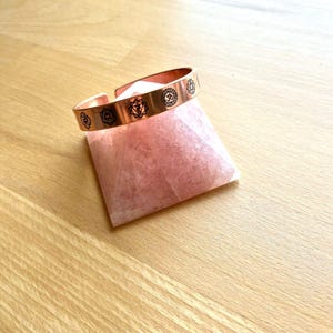 May include: A copper-colored cuff bracelet with etched chakra symbols rests on a pink crystal pyramid. The bracelet is open and adjustable. The background is a light-colored wooden surface.