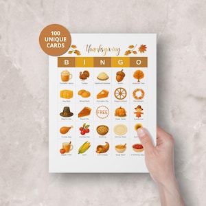 May include: Thanksgiving Bingo game card with colorful illustrations of fall-themed items like turkey, pumpkin pie, and cornucopia. The card has the text "Thanksgiving" and "100 UNIQUE CARDS". The card is held by a hand.