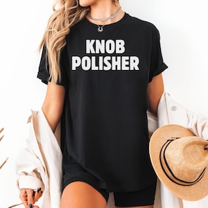 May include: Black t-shirt with the text "KNOB POLISHER" in bold white letters. The shirt is a crew neck style. A straw hat and a white jacket are also visible.