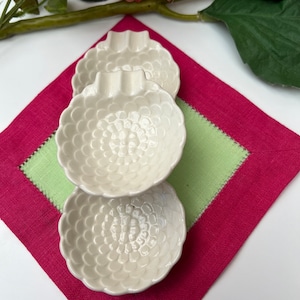 May include: Three white ceramic dishes with a textured, flower-like design. The dishes are stacked on top of each other and are sitting on a green and pink fabric.