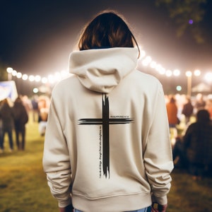 Könnte beinhalten: Ein cremefarbener Kapuzenpullover mit einem großen schwarzen Kreuzdesign auf dem Rücken. Das Kreuz ist mit Pinselstrichen gestaltet, und der Text "PHILIPPIANS 4:13" und "I can do all things through Christ who strengthens me" sind auf der Rückseite aufgedruckt.