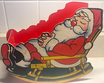 Op de afbeelding: Een vintage kerstdecoratie met een slapende kerstman in een schommelstoel. De kerstman heeft een witte baard, een rood pak en een roze neus. De stoel is rood en groen met gouden accenten, wat een feestelijke vakantiescène creëert.