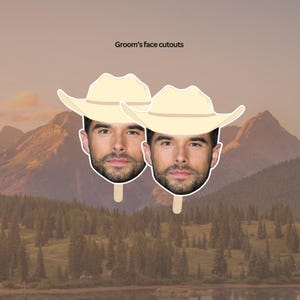 May include: Two cardboard cutouts of a man's face wearing a tan cowboy hat. The cutouts are on sticks and are in front of a mountain landscape.
