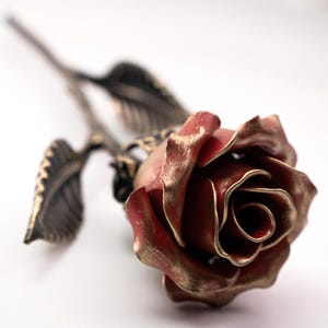May include: A detailed metal rose sculpture, finished in red and gold. The rose has intricate petal and leaf detailing, with a long stem. The metalwork has a vintage, antique look, suitable for home decor or as a unique gift.