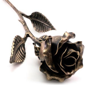 May include: A metal rose sculpture featuring a dark, burnished bronze finish with lighter highlights on the edges of the petals and leaves. The stem is slender and textured, supporting a single bloom with layered, curved petals and two leaves.