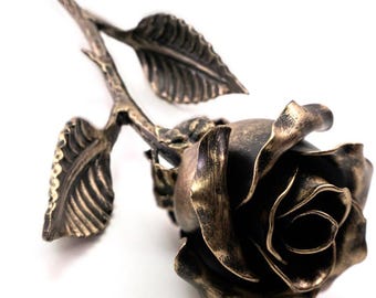 May include: A metal rose sculpture featuring a dark, burnished bronze finish with lighter highlights on the edges of the petals and leaves. The stem is slender and textured, supporting a single bloom with layered, curved petals and two leaves.