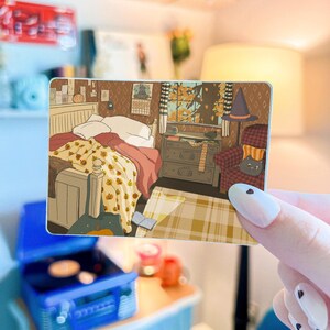 May include: A rectangular sticker featuring an illustration of a cozy bedroom scene. The artwork depicts a bed with a pumpkin-patterned blanket, a window with autumn leaves, and a cat in a witch's hat. A blue record player is in the foreground.
