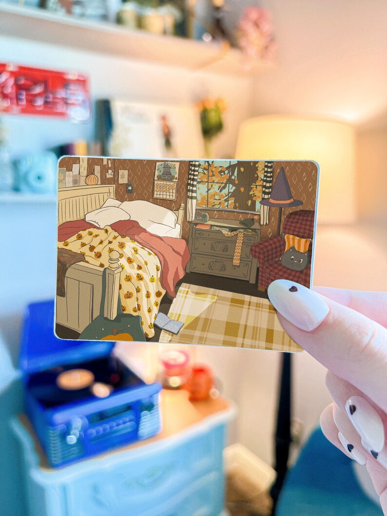 May include: A rectangular sticker featuring an illustration of a cozy bedroom scene. The artwork depicts a bed with a pumpkin-patterned blanket, a window with autumn leaves, and a cat in a witch's hat. A blue record player is in the foreground.