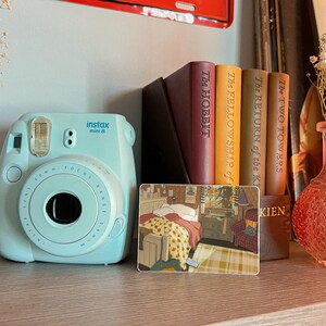 May include: A light blue Instax Mini 8 instant camera sits on a wooden shelf next to a stack of books, including "The Hobbit" and "The Fellowship of the Ring." A decorative card with a room scene and a pink glass vase with dried flowers are also present.