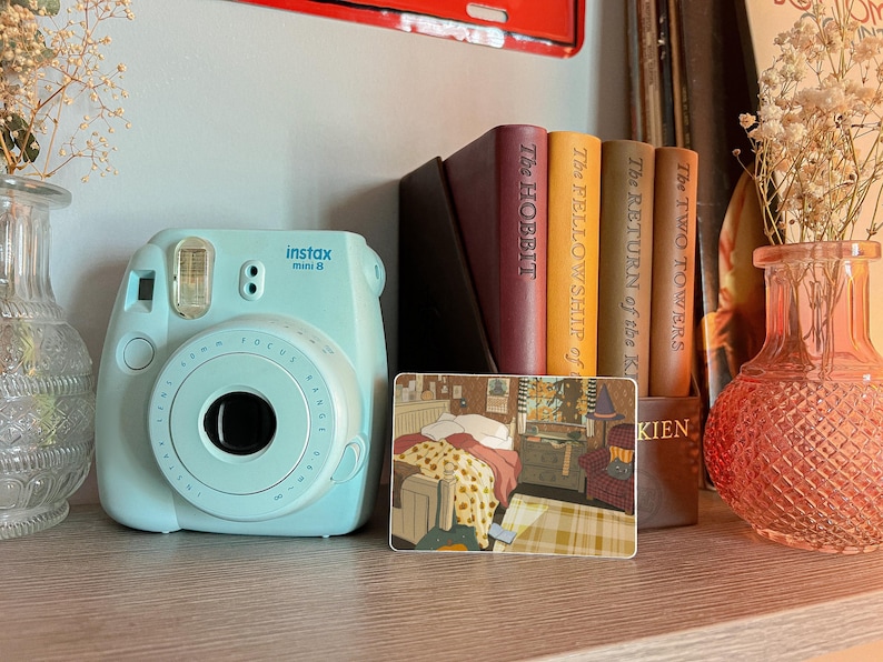 May include: A light blue Instax Mini 8 instant camera sits on a wooden shelf next to a stack of books, including "The Hobbit" and "The Fellowship of the Ring." A decorative card with a room scene and a pink glass vase with dried flowers are also present.