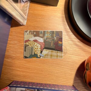 May include: A rectangular sticker featuring an illustration of a cozy bedroom scene. The artwork depicts a bed with a floral blanket, a dresser, a window with a view, and a patterned armchair. The sticker is placed on a wooden surface.