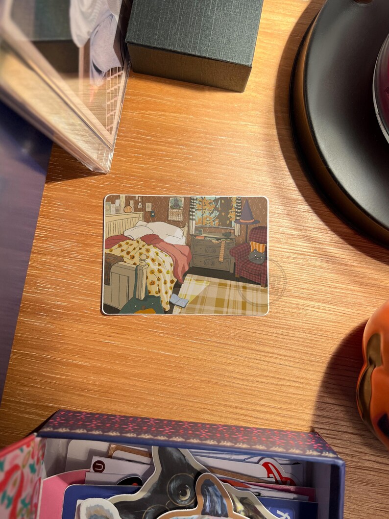 May include: A rectangular sticker featuring an illustration of a cozy bedroom scene. The artwork depicts a bed with a floral blanket, a dresser, a window with a view, and a patterned armchair. The sticker is placed on a wooden surface.
